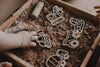 A child's hand reaches into a wooden box filled with sand, exploring the Construction Cutter Set - Eco Playdough Tool made from wood. Another hand is barely visible on the right edge of the image, capturing a moment of sensory play.