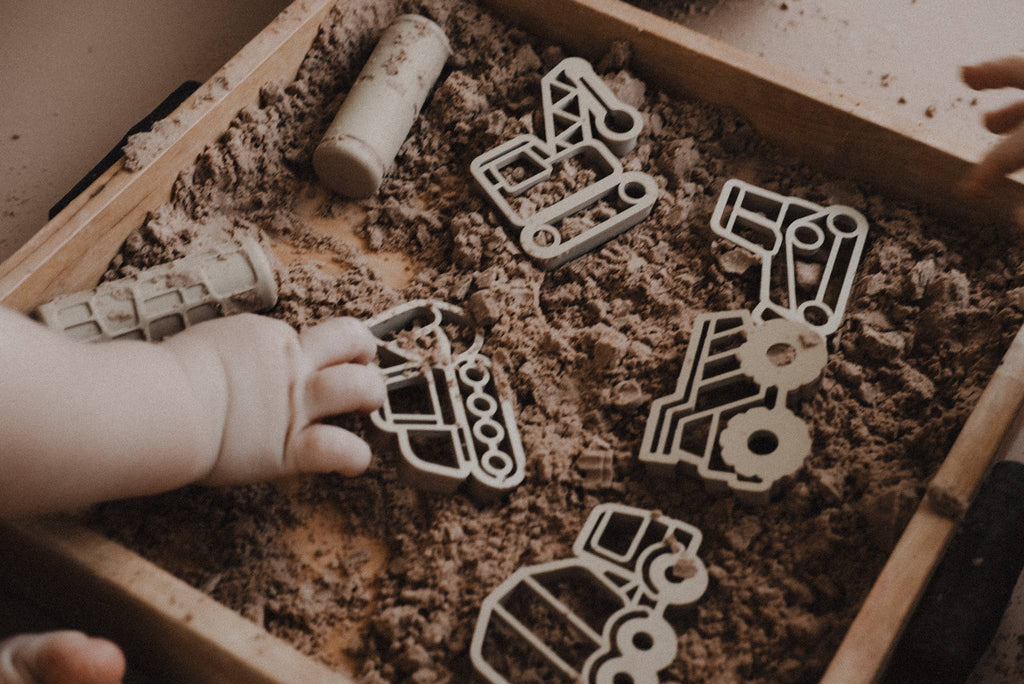 A child's hand reaches into a wooden box filled with sand, exploring the Construction Cutter Set - Eco Playdough Tool made from wood. Another hand is barely visible on the right edge of the image, capturing a moment of sensory play.