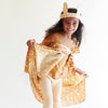 A smiling child in a tan shirt, cream pants, and Sarah's Silks Animal Print Playsilk - Fawn cape with white spots and matching deer ear headband stands against a plain white background, holding out the cape’s sides.