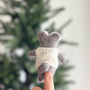 A Baby Koala Finger Puppet/Ornament in gray felt, wearing a white knitted sweater, is perched on a fingertip with a blurred green tree in the background.
