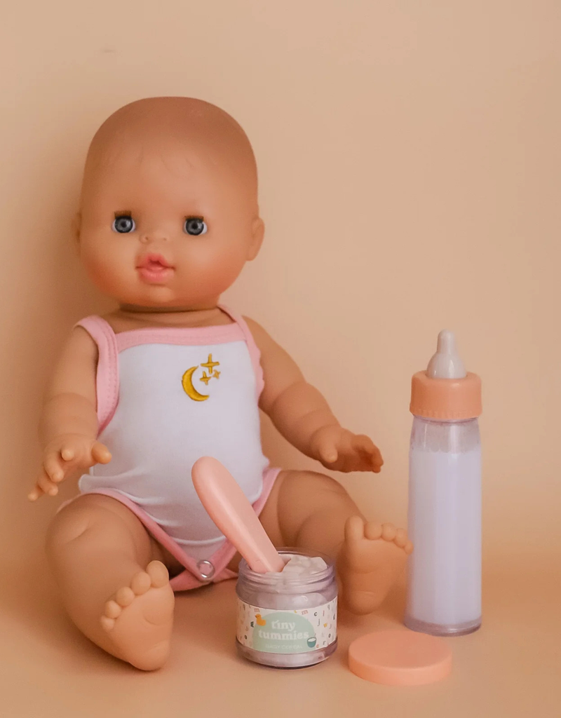 A baby doll wearing a white onesie adorned with a yellow crescent moon sits against a beige background. In front, the "My First Tiny Tummies Gift Set - Milk and Cereal" food jar and a baby bottle create an enchanting scene for little imaginations.