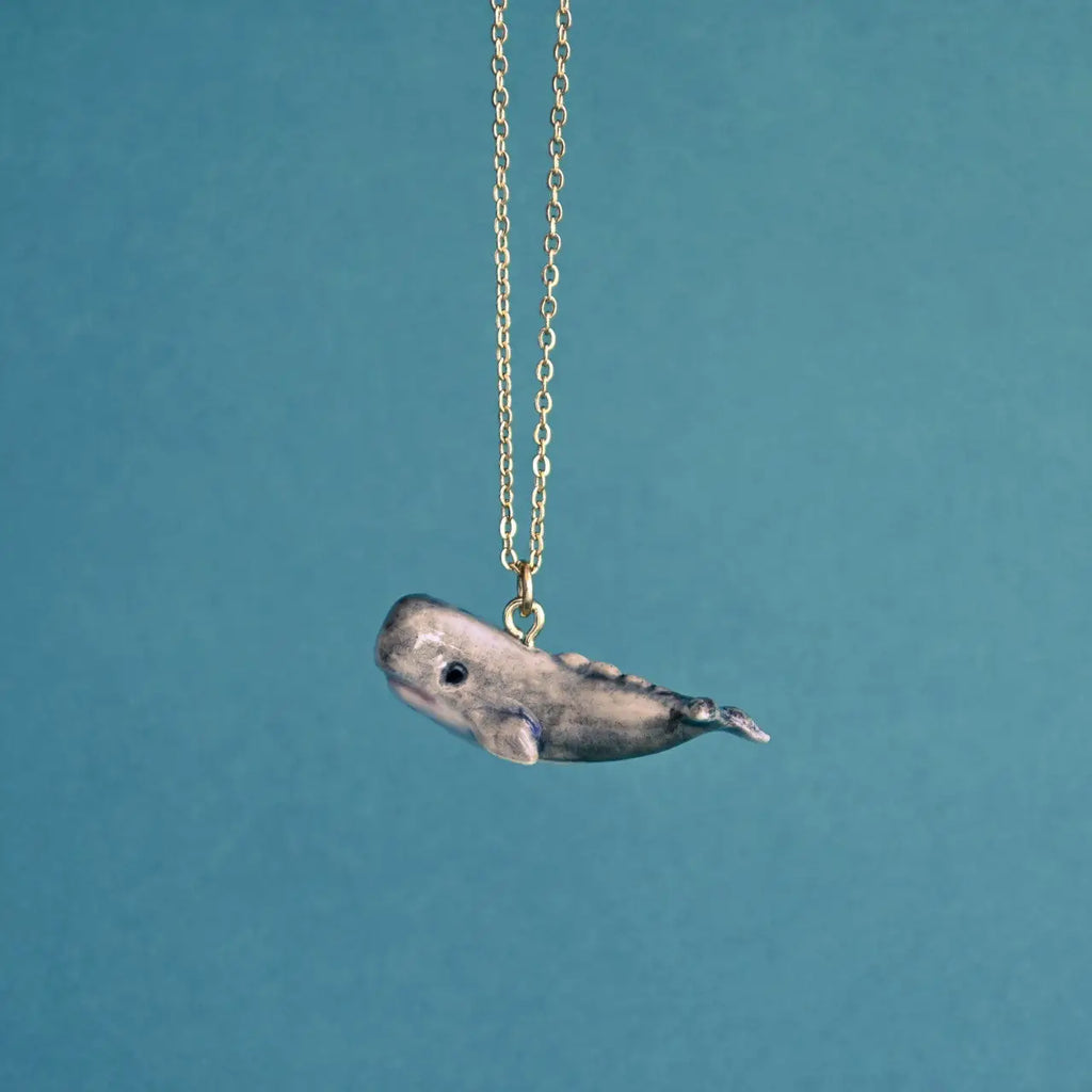 A minimalist pendant shaped like a Whale Necklace, hand painted and hanging from a delicate gold chain, against a muted blue background.