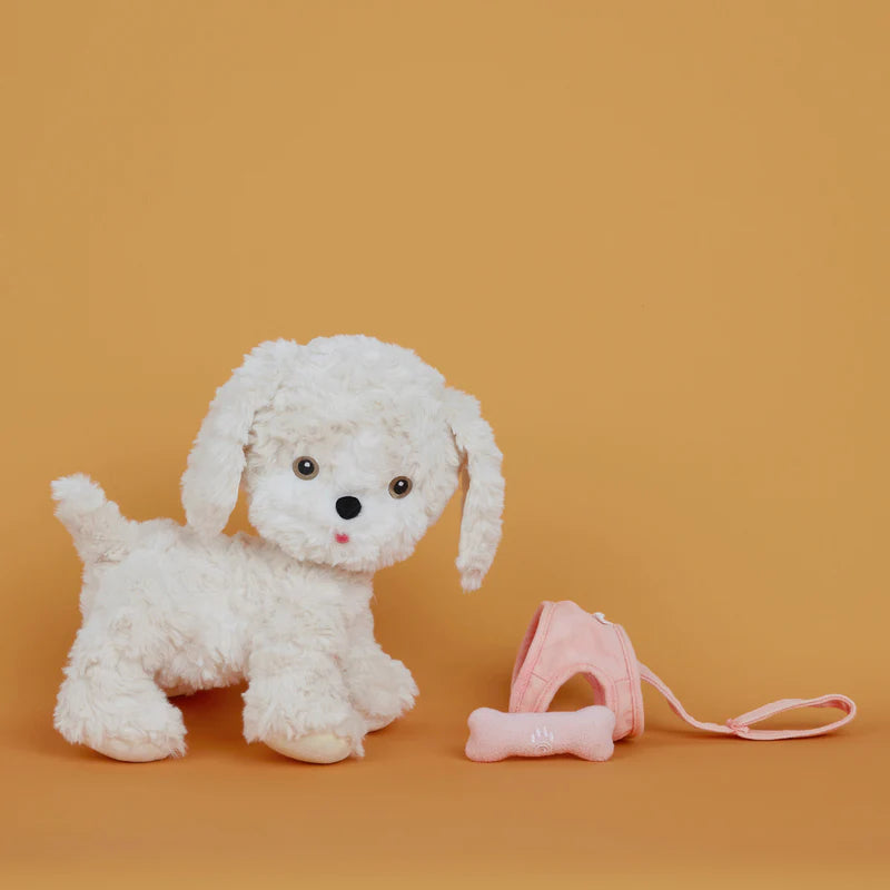 A plush white Olli Ella Dinkum Dog with black eyes and a red nose, sitting next to a pink face mask and a small magnetic bone accessory, against a plain orange background.