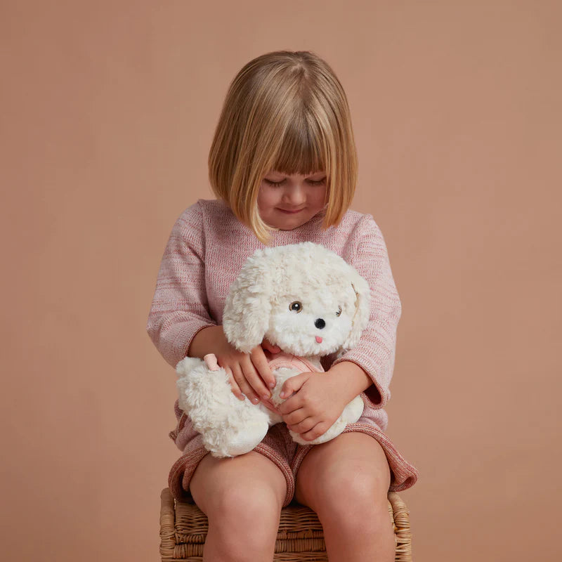 A young girl with a bob haircut, wearing a pink sweater, sits on a wicker stool hugging a fluffy white Olli Ella Dinkum Dog with a magnetic bone accessory, looking down with a gentle smile.