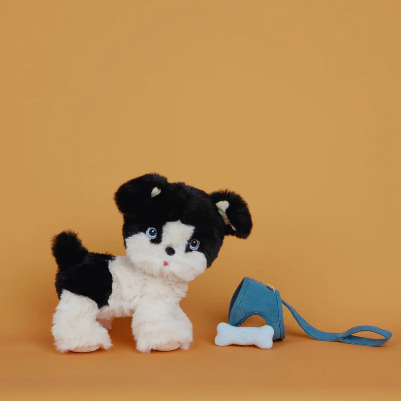A plush toy Olli Ella Dinkum Dog with black and white fur, featuring big round eyes and a small red tongue, stands next to a tiny blue backpack against a plain orange background. The Dinkum Dogs figure includes