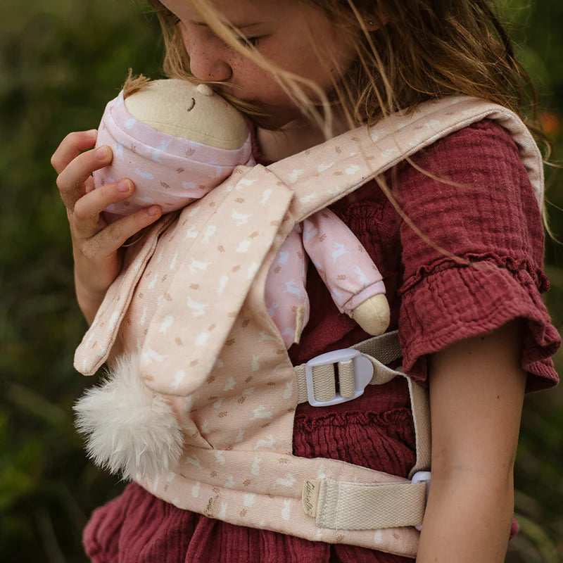 A young girl lovingly kisses a doll wrapped in a pink, star-patterned swaddle, which she carries in an Olli Ella x Odin Parker Dinkum Dolls Cottontail Carrier – Lapin. The focus is on the cudd