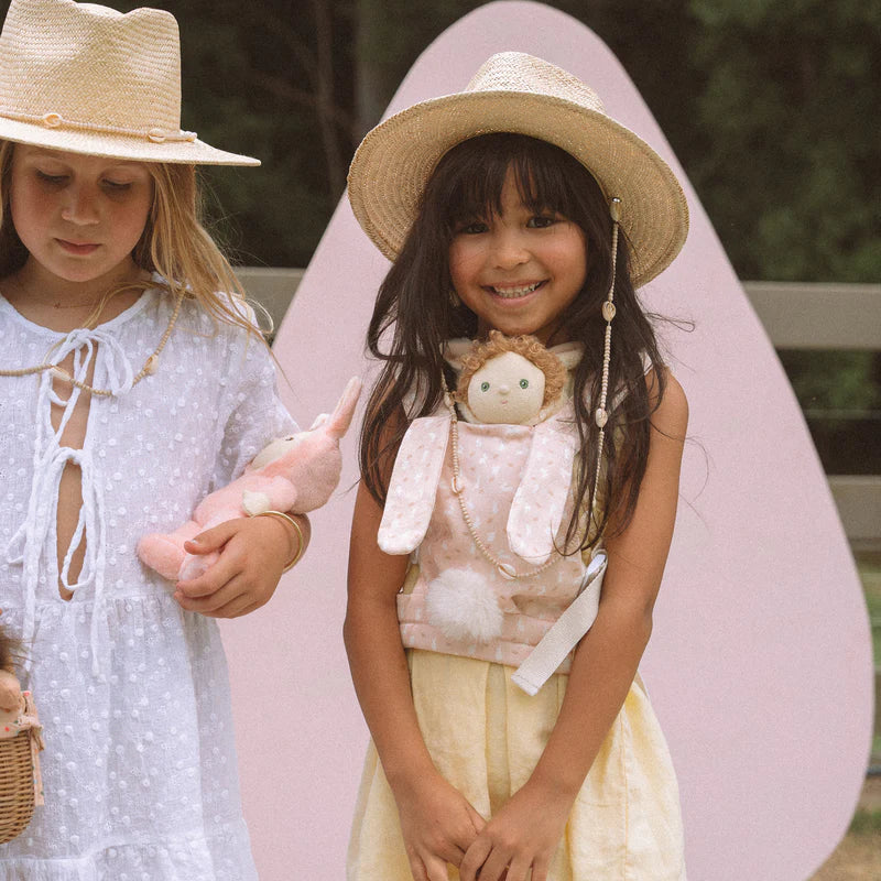 Two young girls in straw hats holding Olli Ella x Odin Parker Dinkum Dolls Cottontail Carrier – Lapin plush toys, one smiling broadly at the camera, with a soft focus background of greenery.