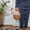 A toddler stands holding a small Easter basket with an Olli Ella | Dinky Dinkums - Bella Bunny inside. The setting includes a woven rug and flowers beside them, emphasizing a cozy, indoor environment.