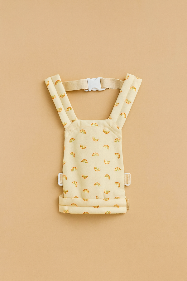 The Olli Ella Doll Carrier, a small yellow mini carrier with a vibrant rainbow print and adjustable straps, is shown on a solid beige background.