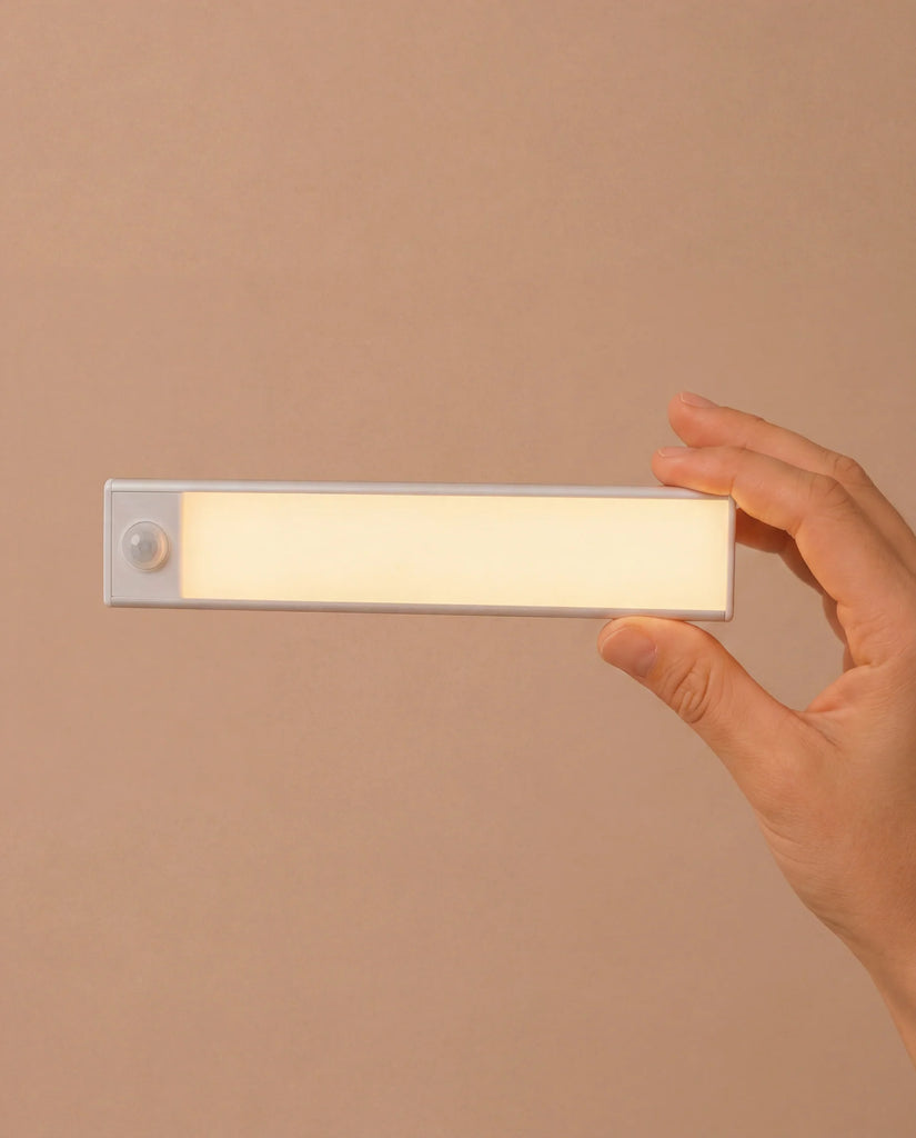 A hand holds a rectangular, battery-powered LED light with a frosted cover and a small round sensor on one end, against a plain beige background—similar to the modern lighting featured in the Fully Furnished Dollhouse With Lights, House of New York - The Six-Storey Giant.