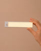 A hand holds a rectangular, slim, white LED light with a small circular sensor on one end, set against a plain beige background—an ideal accessory for illuminating every room in your House of Paris Luxury Dollhouse – A Tale of Parisian Magic, or for bringing a touch of modern elegance to this enchanting Parisian miniature home.