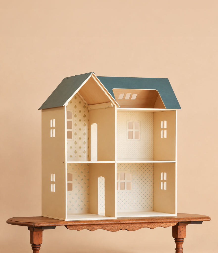 A Maileg Dollhouse, House of Miniature with a blue roof stands on a wooden table. This charming dollhouse features four rooms, wallpapered interiors, and cut-out windows and doors, making it perfect for arranging your favorite Maileg furniture accessories. The background is a plain light beige color.
