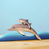 Two wooden dolphins from the Bumbu Handmade Wooden Dolphin Family Set leap over stylized blue waves on a wooden surface with a light blue background.