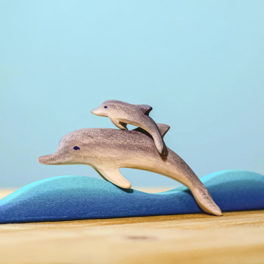 Two wooden dolphins from the Bumbu Handmade Wooden Dolphin Family Set leap over stylized blue waves on a wooden surface with a light blue background.