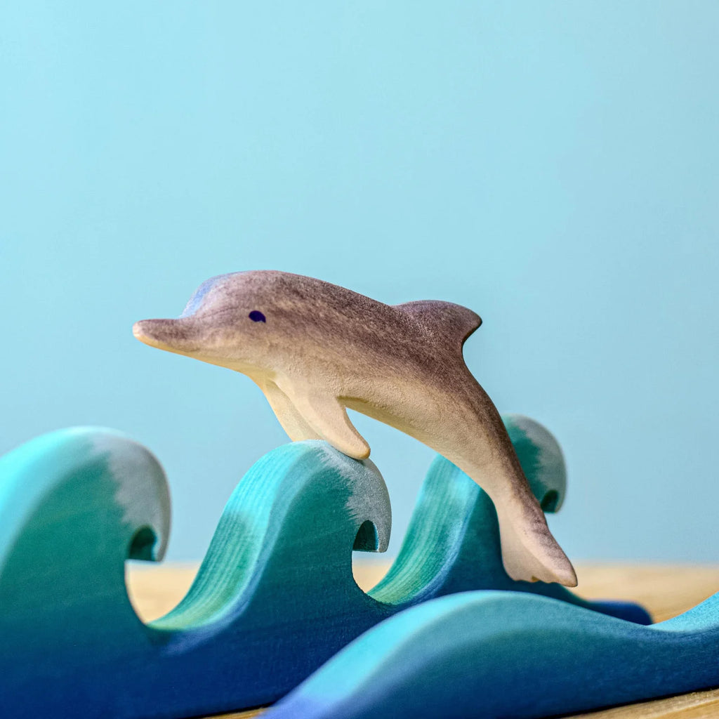 A handcrafted wooden dolphin leaps over eco-friendly blue waves on a light blue background—a charming addition to any Bumbu Handmade Wooden Dolphin Family Set.