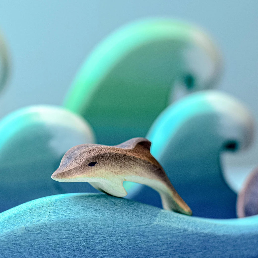 The Bumbu Handmade Wooden Dolphin Family Set features handcrafted dolphins nestled among painted blue and green waves, creating an eco-friendly ocean scene.