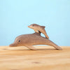 Two dolphins from the Bumbu Handmade Wooden Dolphin Family Set are arranged so the smaller one rides on the larger’s back, set against a light blue background with a wooden surface below.