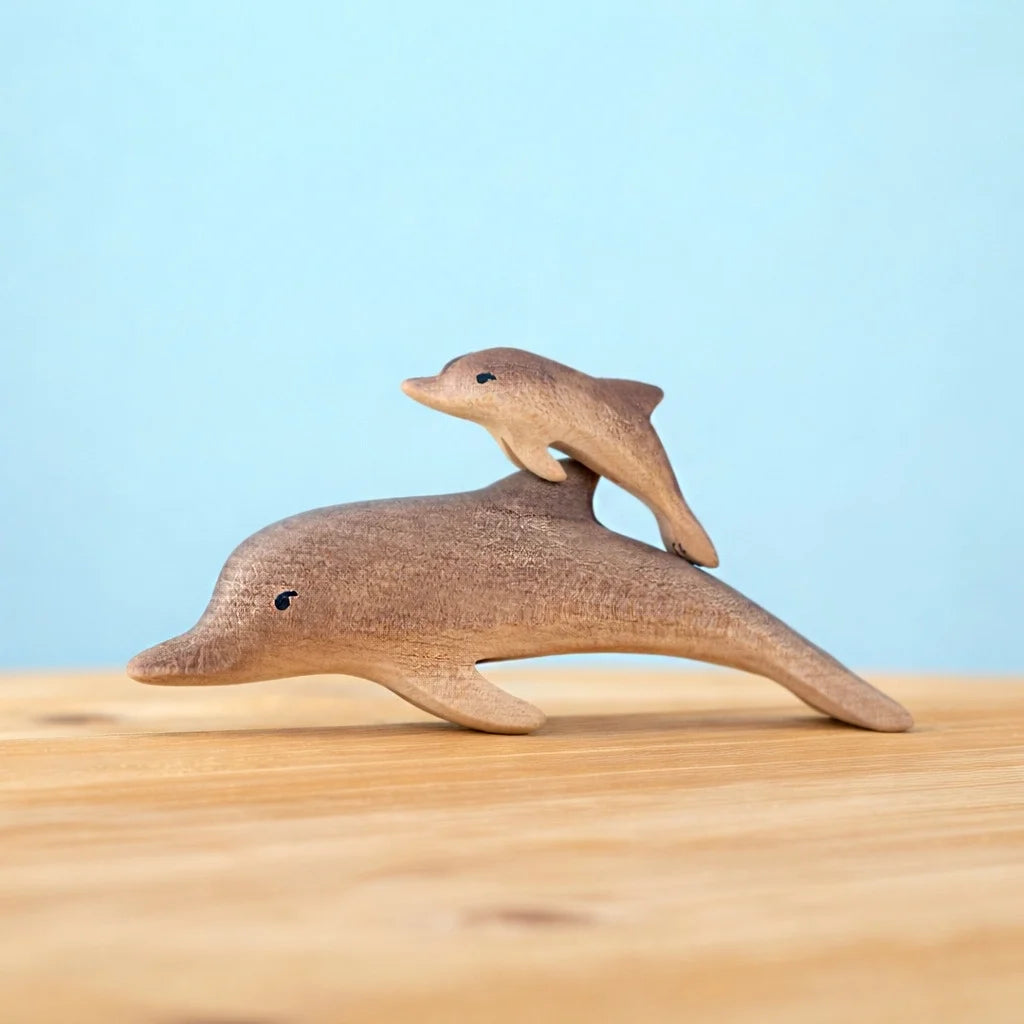 Two dolphins from the Bumbu Handmade Wooden Dolphin Family Set are arranged so the smaller one rides on the larger’s back, set against a light blue background with a wooden surface below.