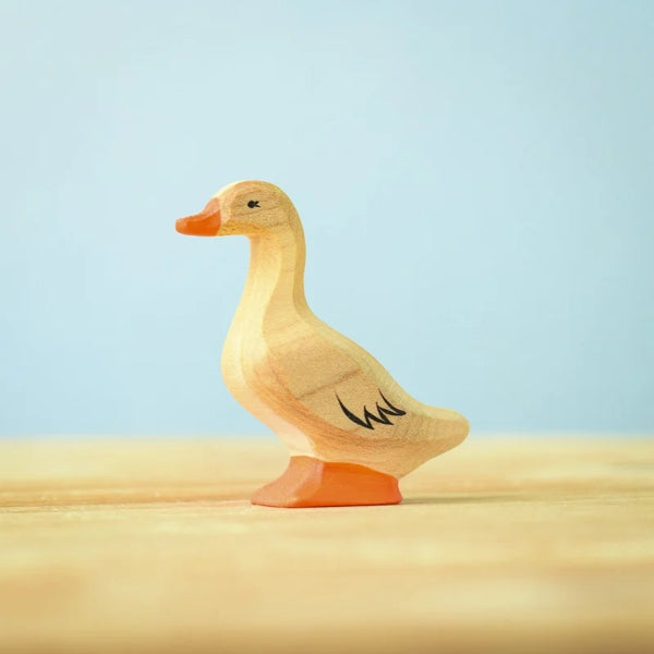 The Bumbu Handmade Wooden Domestic Duck features a small, handcrafted design with an orange beak and feet, black detailing on the eyes and wing, standing on a light wooden surface against a pale blue background.