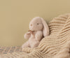 The Maileg Plush Bunny, Mini - Light Powder, a soft stuffed animal with floppy ears and a brown ribbon, sits on a textured beige knit blanket atop a patterned surface. It’s crafted from recycled polyester with a light tan wall as the backdrop.