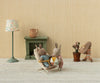 Three Maileg Rabbit With Accessories, Micro - Sand - Rose sit in a cozy room: two read together on a lounge chair while the third waters a plant. A green mantel, lamp, and jug complete the sweet scene against beige wallpaper.