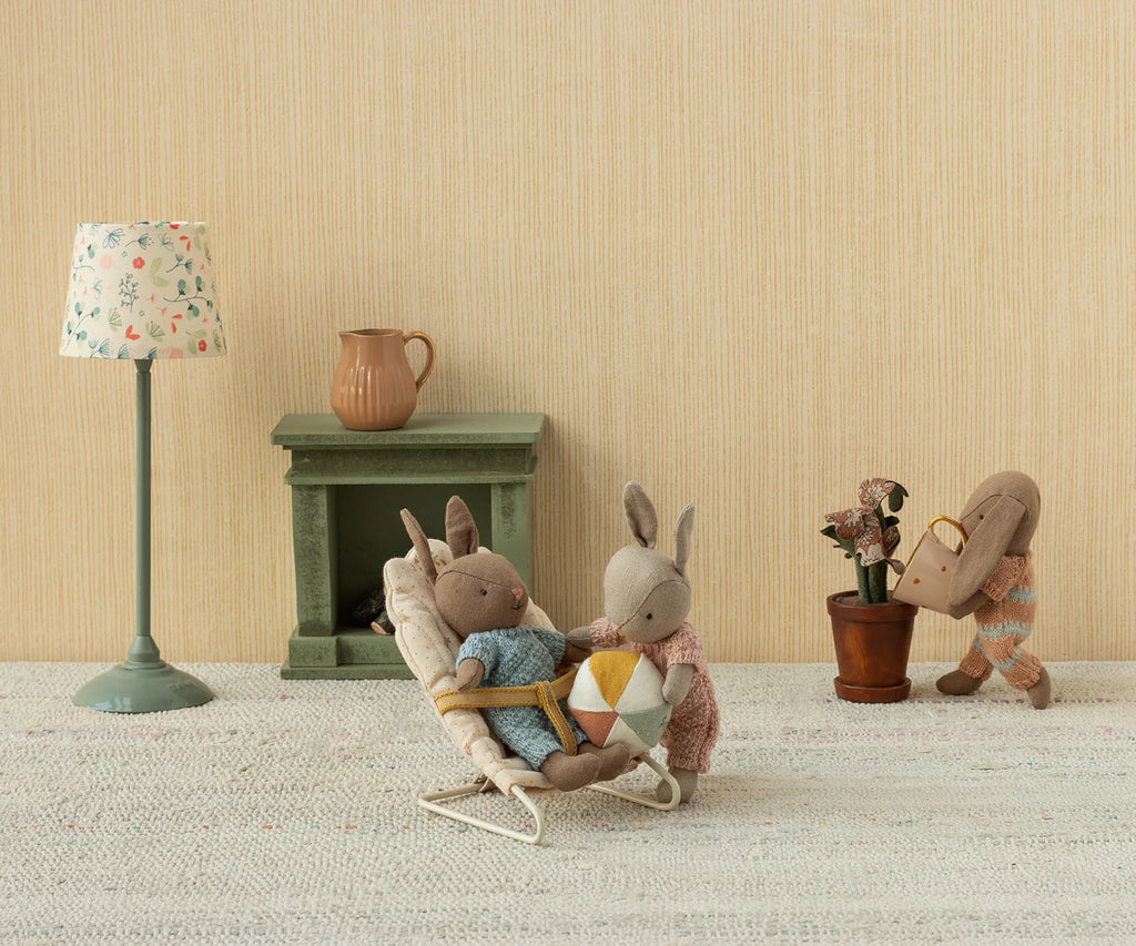 Three Maileg Rabbit With Accessories, Micro - Sand - Rose sit in a cozy room: two read together on a lounge chair while the third waters a plant. A green mantel, lamp, and jug complete the sweet scene against beige wallpaper.