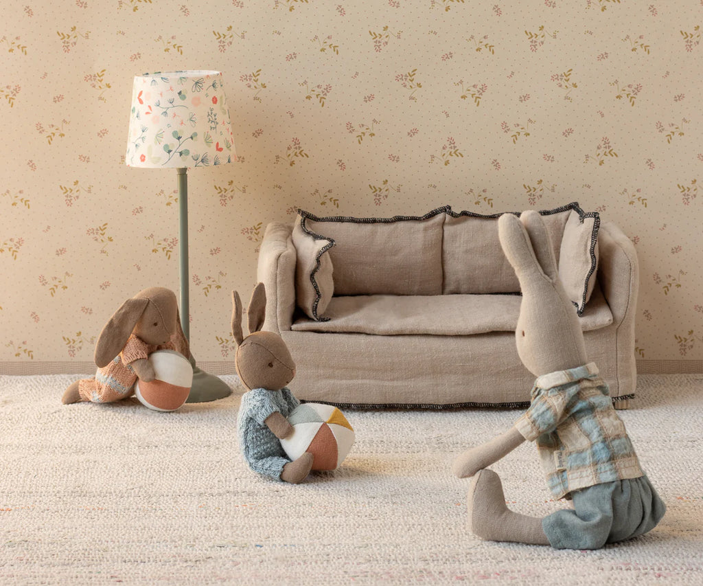 The Maileg Micro Bunny With Ball - Brown - Blue (with magnetic hands) plays with a soft ball in a cozy room featuring a beige sofa, floor lamp, and floral wallpaper, all set in gentle pastel tones.