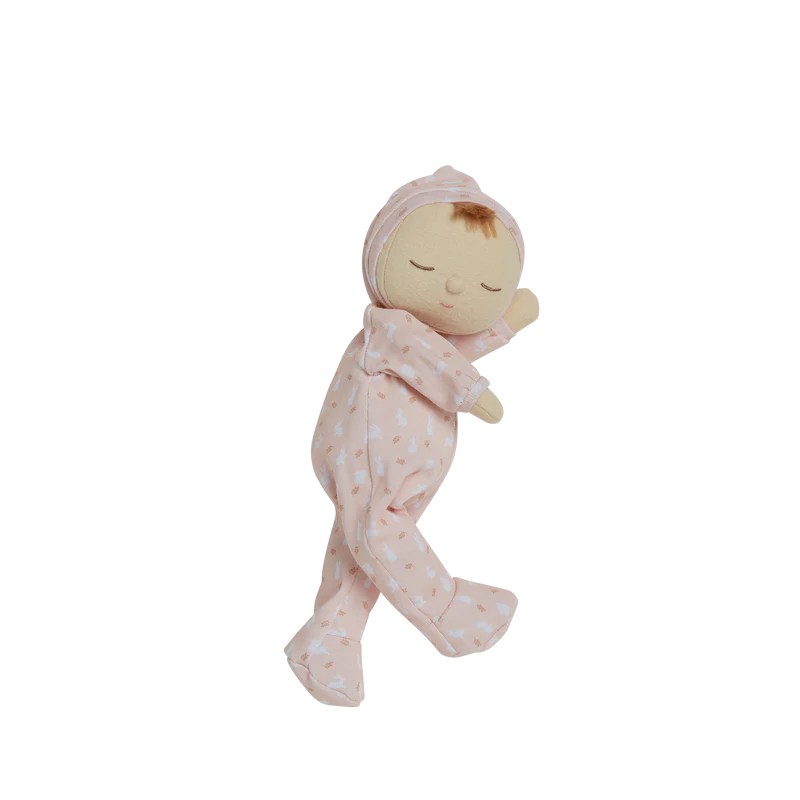 A plush doll from the Olli Ella x Odin Parker Dozy Dinkums - Blossom line depicting a sleeping baby swaddled in a pink blanket with heart patterns, shown in motion with blurred horizontal lines against a black background.