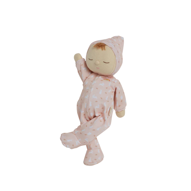 A Olli Ella x Odin Parker Dozy Dinkums - Blossom plush doll dressed in a pink onesie with a hood, decorated with subtle heart patterns, appears to be floating or hanging, against a black background with horizontal white lines.