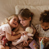 Three young girls lying close together, sharing a moment of joy. Two of them hold Olli Ella x Odin Parker Dozy Dinkums - Blossom dolls while one reads a picture book. They appear content and engaged in their