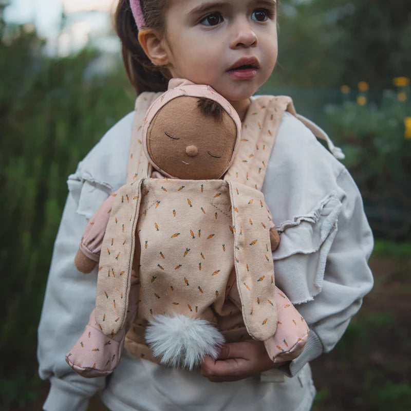 A young child holding an Olli Ella x Odin Parker Dozy Dinkum - Bugsy Hopscotch plush doll with a happy expression, standing outdoors with greenery in the background. The doll features a cute, simplistic face and a fluffy tail.