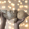 A pair of hands holds up the Olli Ella Dozy Dinkum - Custard Sweetheart doll, dressed in a soft cream outfit adorned with small red hearts, against a backdrop of warm, glowing lights—an adorable creation that makes the perfect newborn gift.