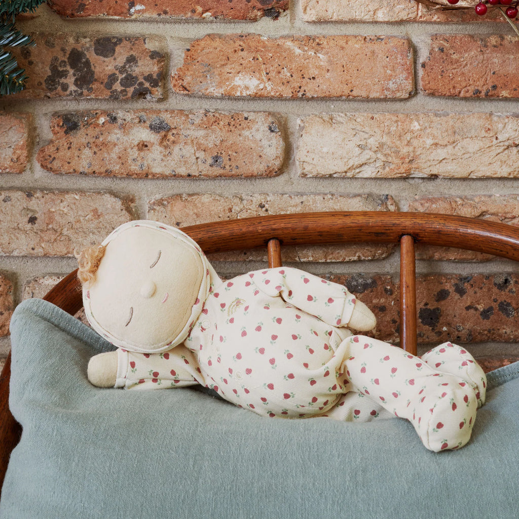 The Olli Ella Dozy Dinkum - Custard Sweetheart, a soft fabric doll dressed in patterned pajamas, rests on a blue cushion against a brick wall—an adorable creation that’s perfect as a newborn gift.