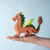 A hand holds the Bumbu Handmade Wooden Dragon, featuring an orange-brown body, green spines, and yellow-green wings against a light blue background.