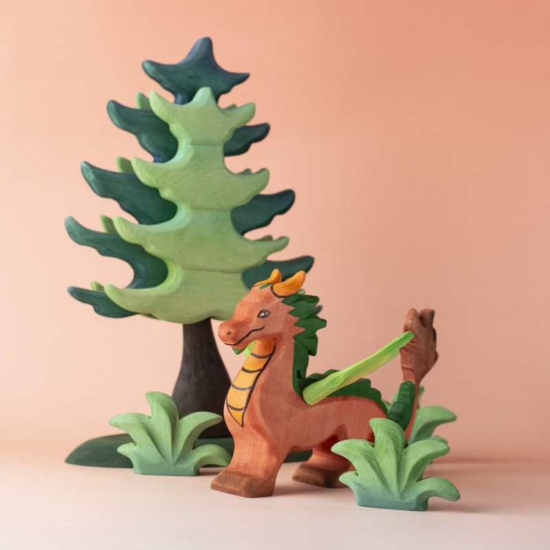The Bumbu Handmade Wooden Dragon stands proudly before a tree and bushes, all lovingly crafted. Set against a soft peach background, this fantasy toy scene invites imaginative play and adventure.