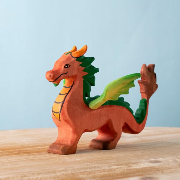 The Bumbu Handmade Wooden Dragon, featuring an orange and yellow body with green wings and accents, stands on a light wooden surface against a plain blue background.