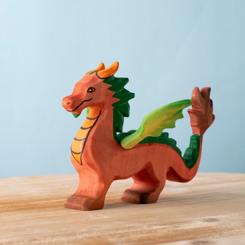 The Bumbu Handmade Wooden Dragon, featuring an orange and yellow body with green wings and accents, stands on a light wooden surface against a plain blue background.