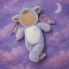 The Olli Ella Cozy Dinkum Dragon - Rory - Lavender, featuring wings, horns, and closed eyes, lies on a purple cloud-patterned blanket with a crescent moon background—ideal for soothing cuddles.