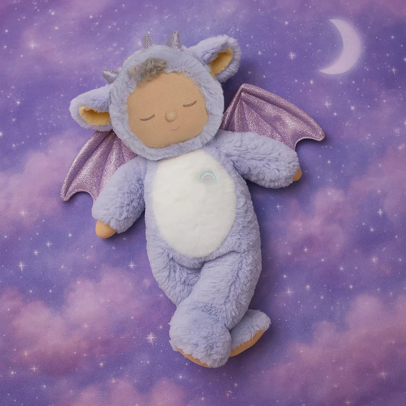The Olli Ella Cozy Dinkum Dragon - Rory - Lavender, featuring wings, horns, and closed eyes, lies on a purple cloud-patterned blanket with a crescent moon background—ideal for soothing cuddles.