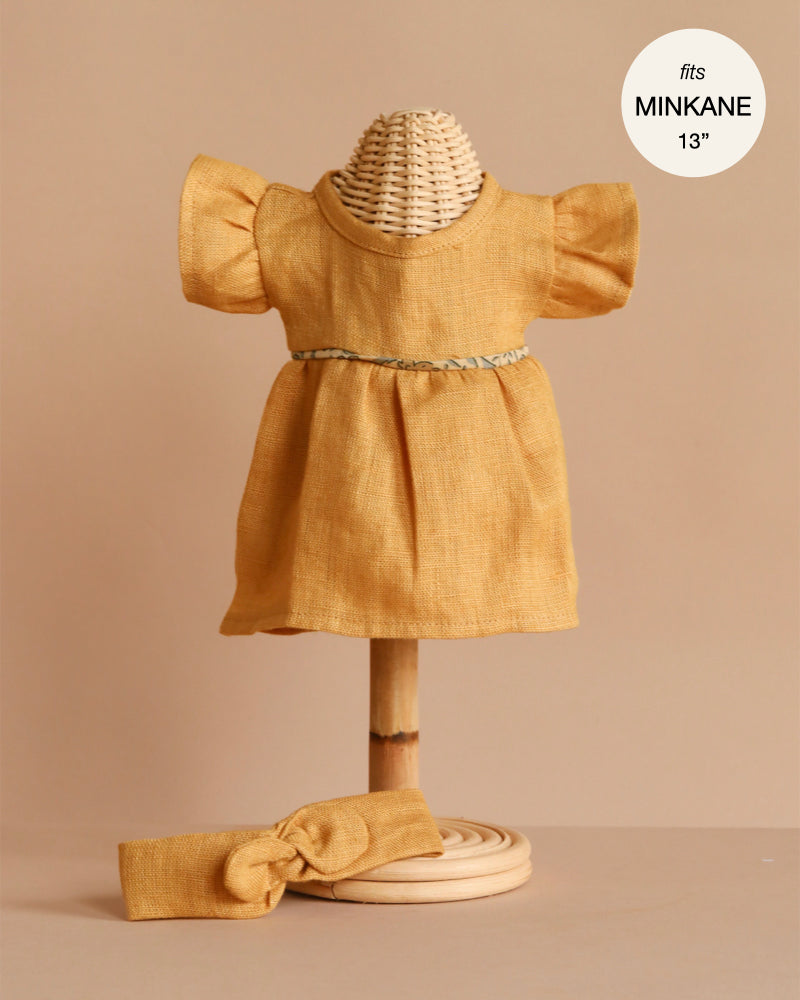The Minikane Doll Clothing Daisy Dress & Headband in Washed Linen - Oatmeal features a short-sleeve dress with ruffled sleeves, braided belt, and matching headband. Ideal for Minikane and other 13” dolls. Text reads "fits MINIKANE 13”.