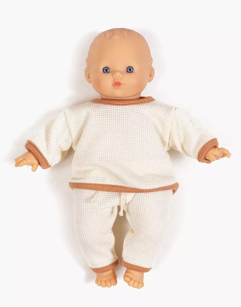 A Minikane Dolls baby with blue eyes wears the Minikane 11" Cocoon Jogger Set in Honeycomb Cotton - Ecru, featuring a cream knit long-sleeve top and matching pants with tan trim, lying on a white background.