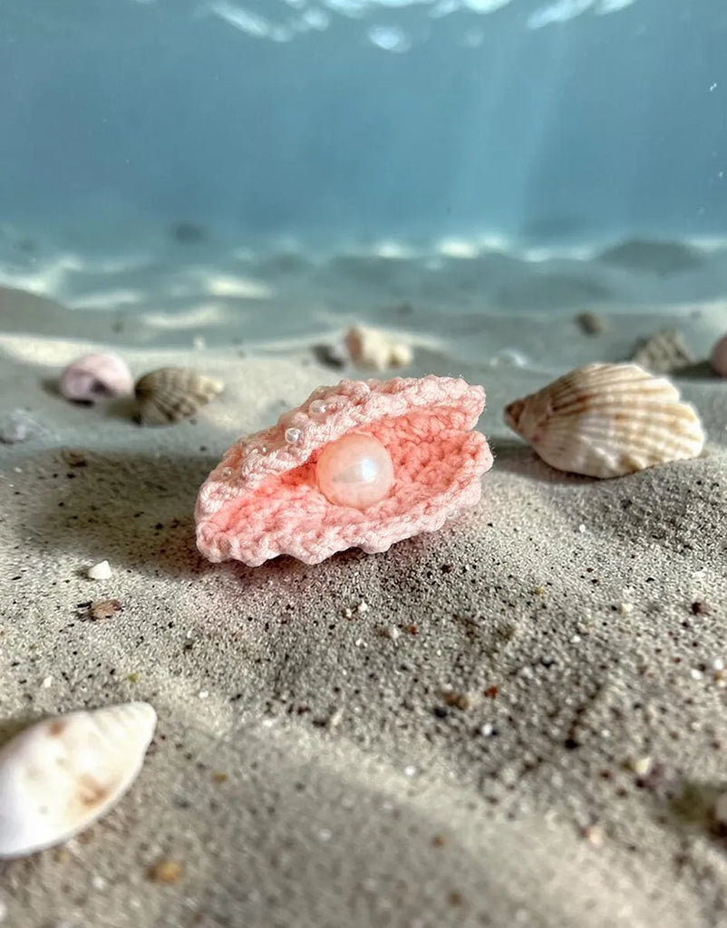 A close-up of the Minikane 13" Doll Clothing | Mermaid Costume & Pink Pearl Seashell Set features a pink crocheted oyster with pearl, seashells on sand, and blue water—ideal for mermaid outfit inspiration or doll accessories.