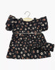 The Minikane Doll Clothing Rylee+Cru Daisy Dress and Headband - Capucine features a black floral dress with puffed short sleeves for 13-inch dolls, shown on a wooden hanger against a white background.
