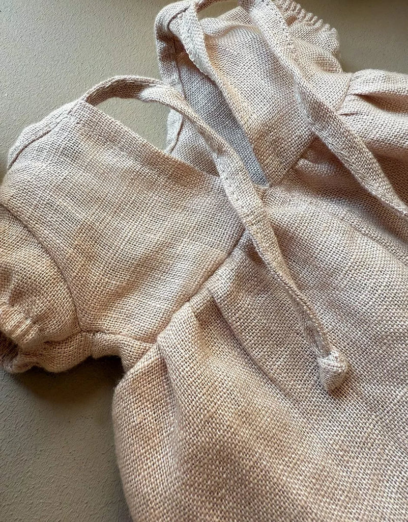 A close-up of the Minikane 11" Doll Clothing Faustine Dress with balloon sleeves in nude linen, designed for Minikane Babies dolls, lying on a neutral surface.