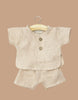 The Minikane 13" Doll Clothing Andrea Short Set in Washed Linen - Nude features a beige short-sleeved top with two wooden buttons and matching shorts, displayed on a small wooden hanger against a plain tan background.