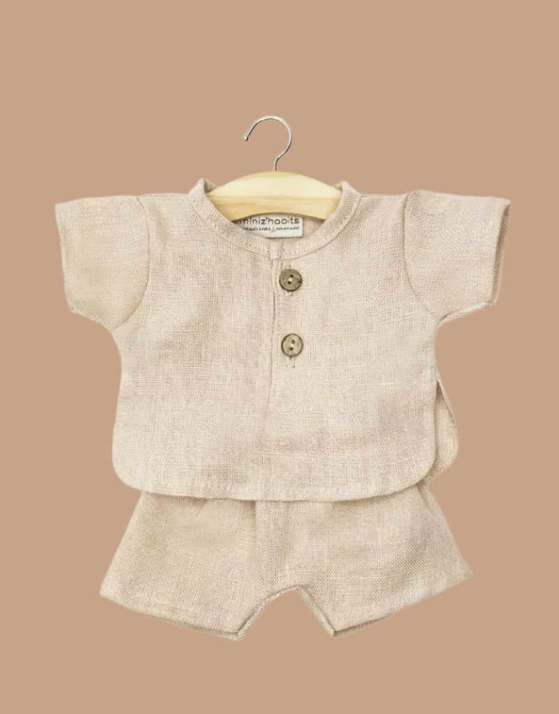 The Minikane 13" Doll Clothing Andrea Short Set in Washed Linen - Nude features a beige short-sleeved top with two wooden buttons and matching shorts, displayed on a small wooden hanger against a plain tan background.