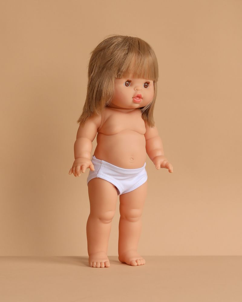 The Minikane Standing Doll (14") - Zoelie features light skin, straight blonde hair with bangs, and white underwear. Anatomically correct and made in Spain, Zoelie stands upright against a plain beige background. Clothing sold separately.