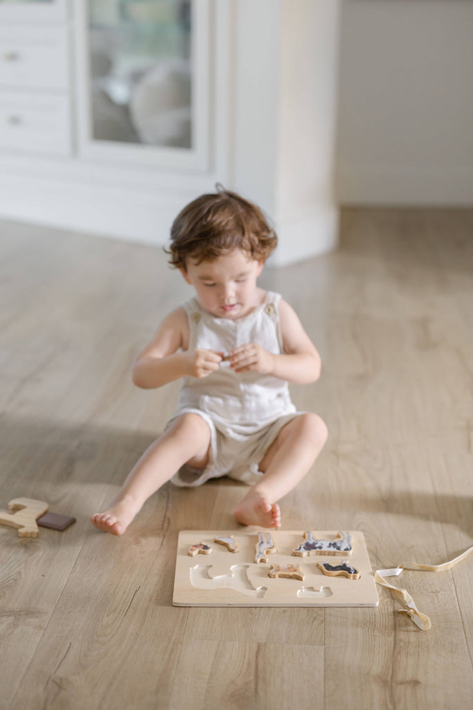 A young child sits on a wooden floor, playing with the Farm Animals Wooden Puzzle: A Tabitha Paige Puzzle, holding a piece and concentrating.