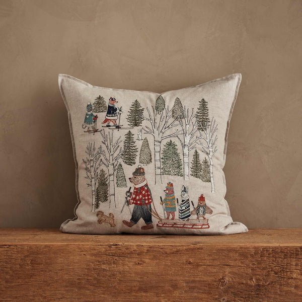 The Coral & Tusk Winter Fun Pillow features woodland animals in scarves and coats skiing, sledding, and walking among snowy trees—perfect holiday decor for your home. Shown on a wooden surface against a tan wall.