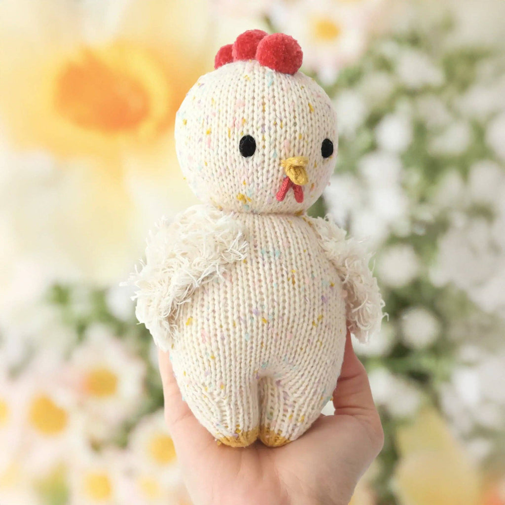A hand holds the Cuddle + Kind Baby Chicken—a cream-colored, hand-knit stuffed animal with a yellow beak, red comb, black eyes, multicolored specks, and fluffy white wings. This heirloom-quality doll is handmade in Peru. Flowers blur in the background.